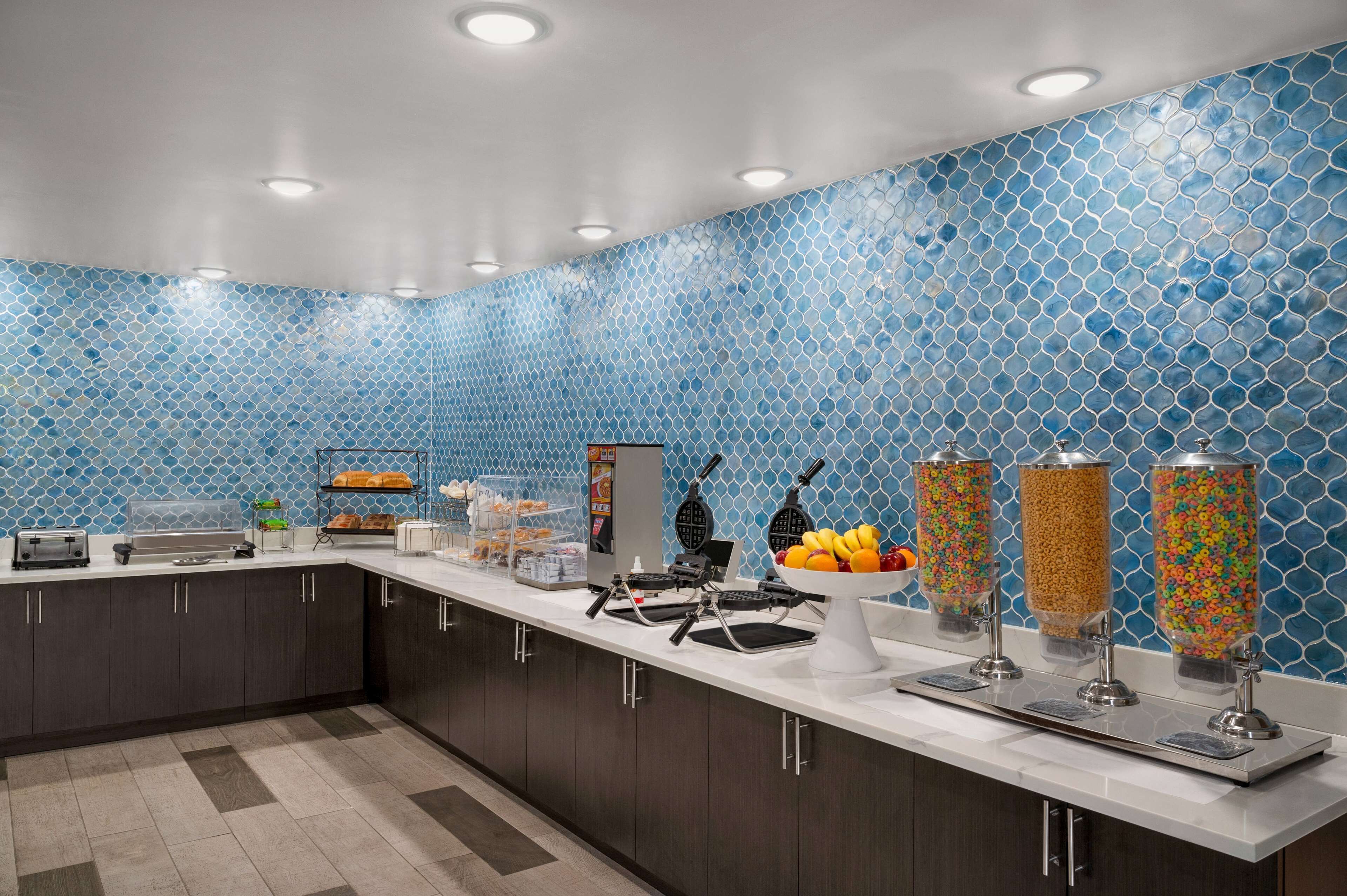 A modern kitchen with a blue mosaic backsplash, a long counter with various appliances, and food dispensers.