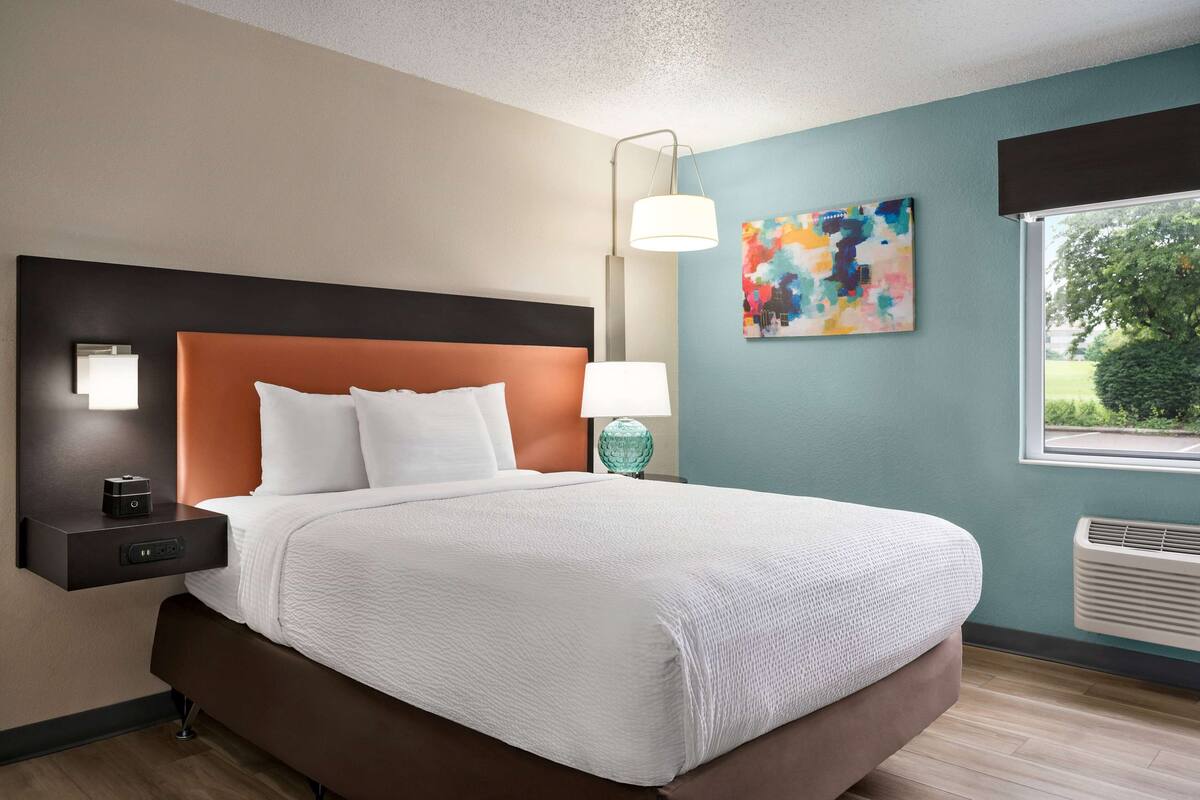 A modern hotel room with a large bed, a nightstand with a lamp, a wall-mounted air conditioner, and a window with a view of greenery.