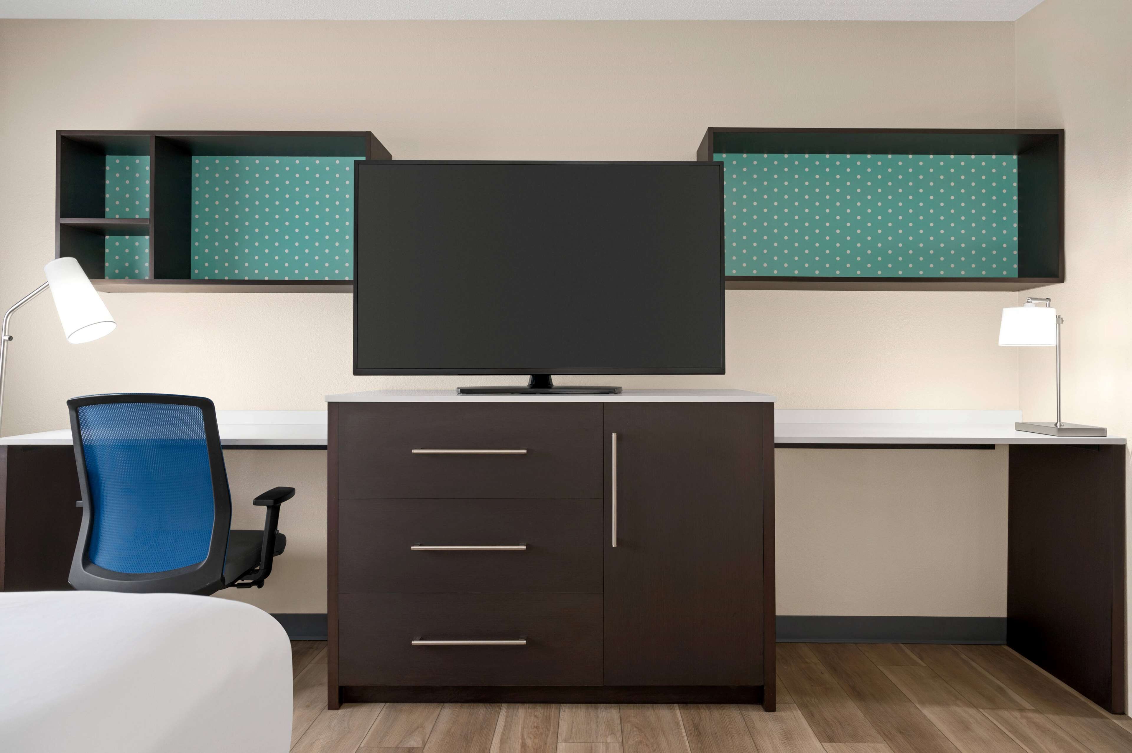 Une chambre d’hôtel moderne avec un bureau en bois foncé, un téléviseur à écran plat, une chaise de bureau bleue et un lit blanc.