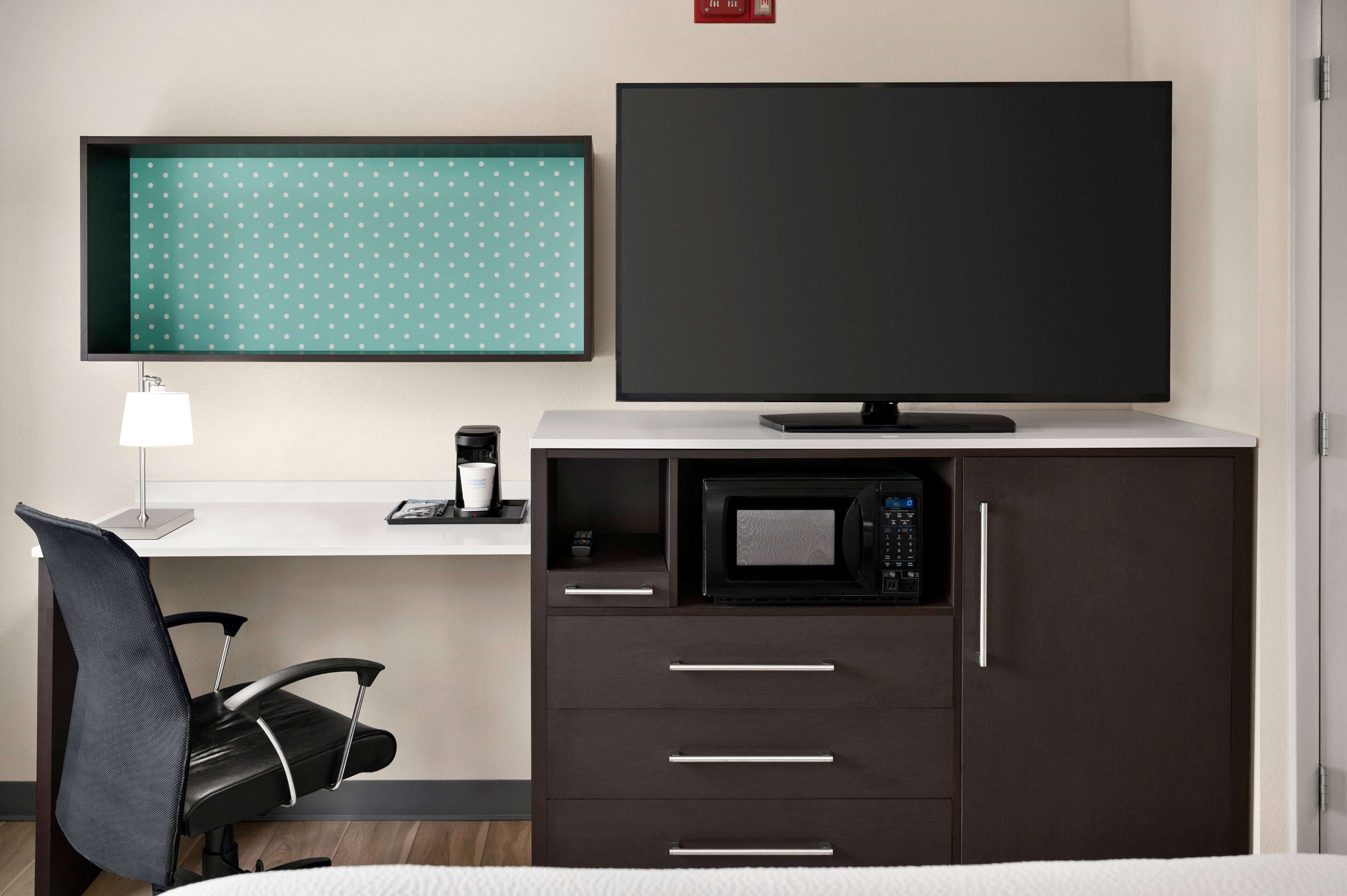 A hotel room with a bed, a desk with a lamp, a chair, a television, a microwave, and a drawer unit.