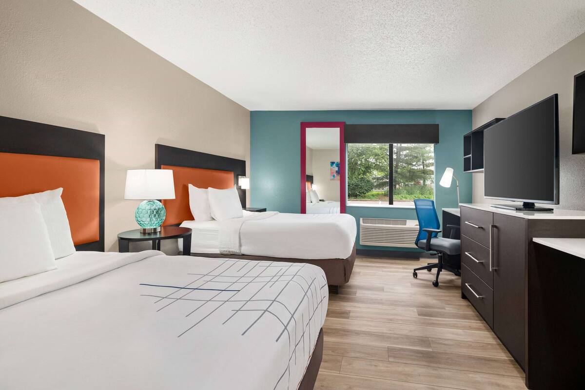 A hotel room with two beds, a desk, a TV, and a view of the outdoors.