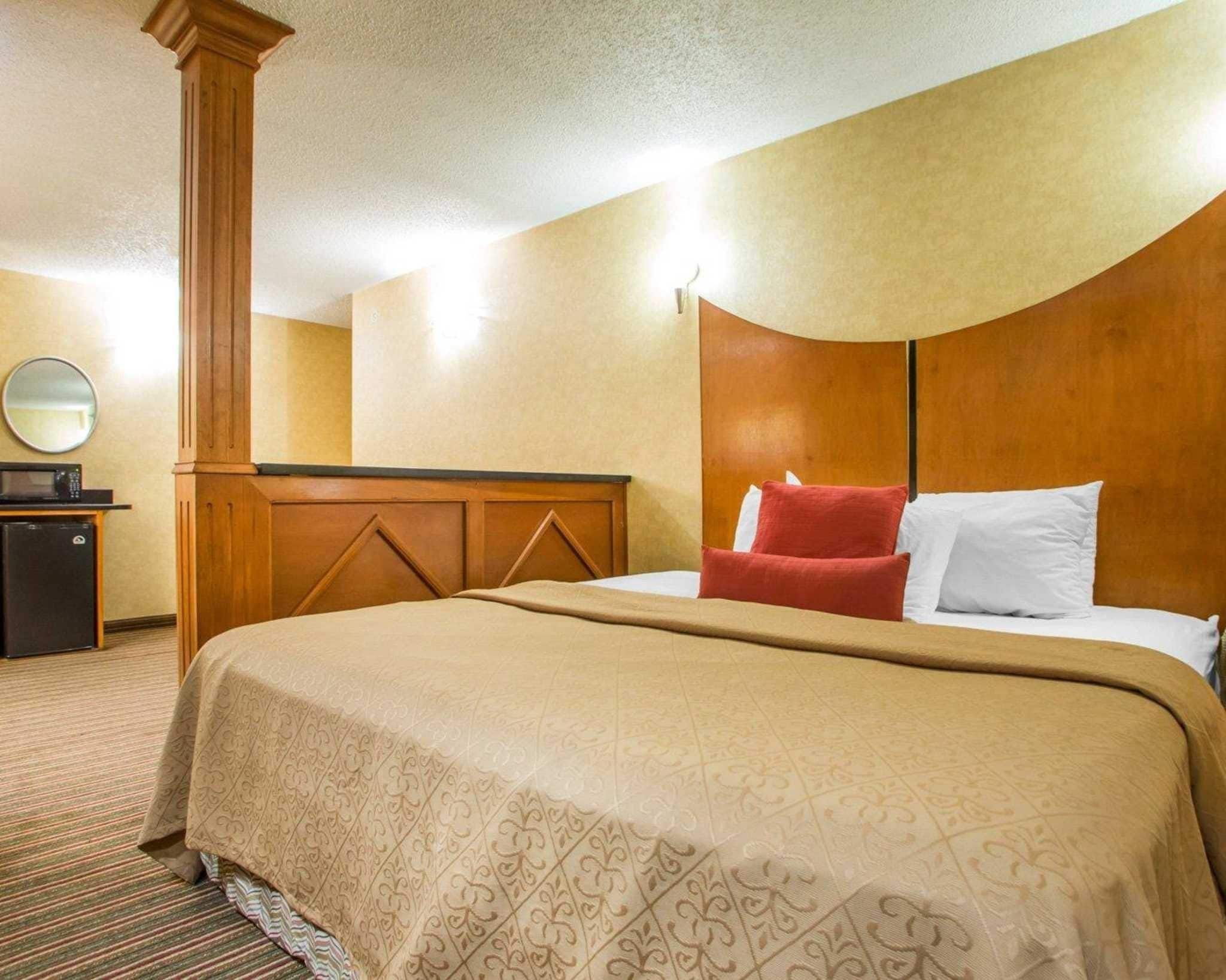 A hotel room with a large bed, a wooden headboard, a desk with a mirror, and a small refrigerator.