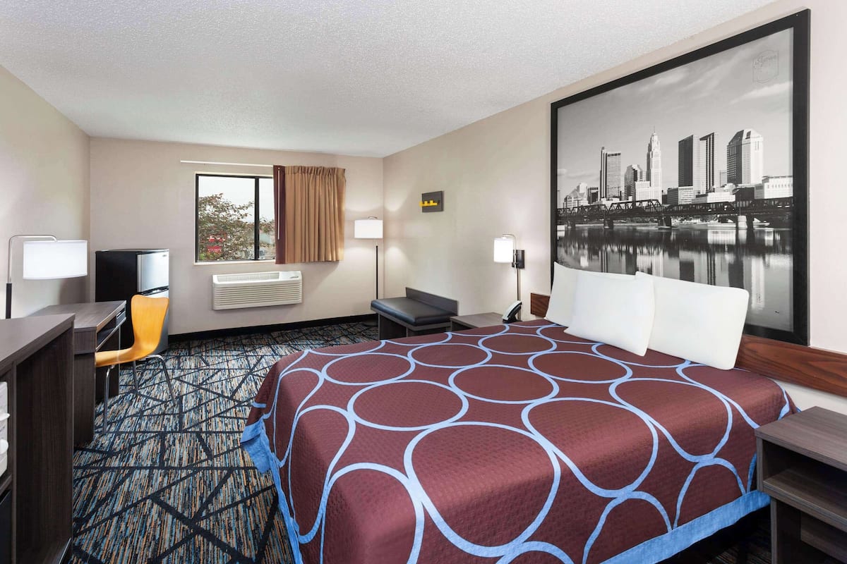 Hotelkamer met een groot bed, een bureau, een stoel, een raam met gordijnen, een luchtverfrisser aan de muur en een stadslandschap schilderij.