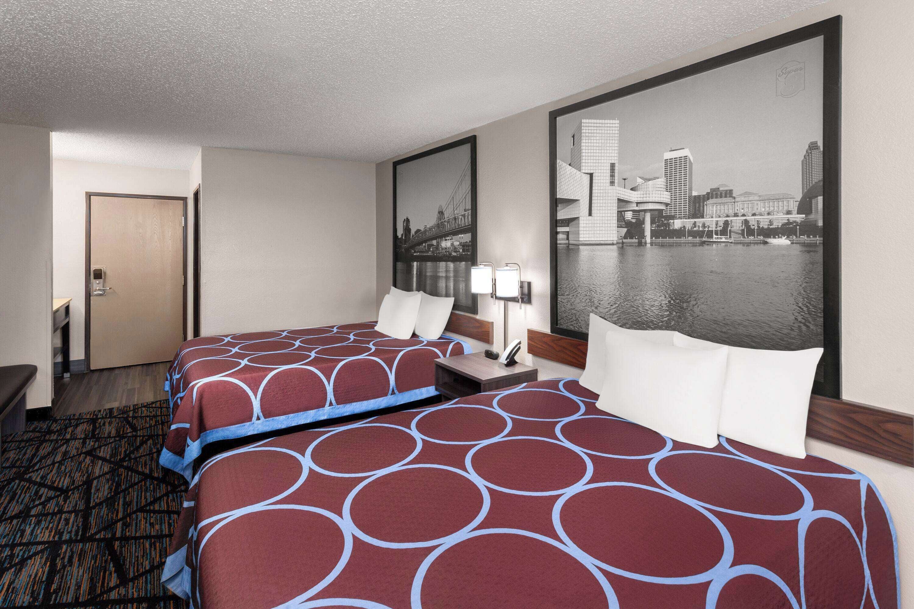 Quarto de hotel com duas camas, vista para a cidade, uma porta e uma pequena mesa com abajur.