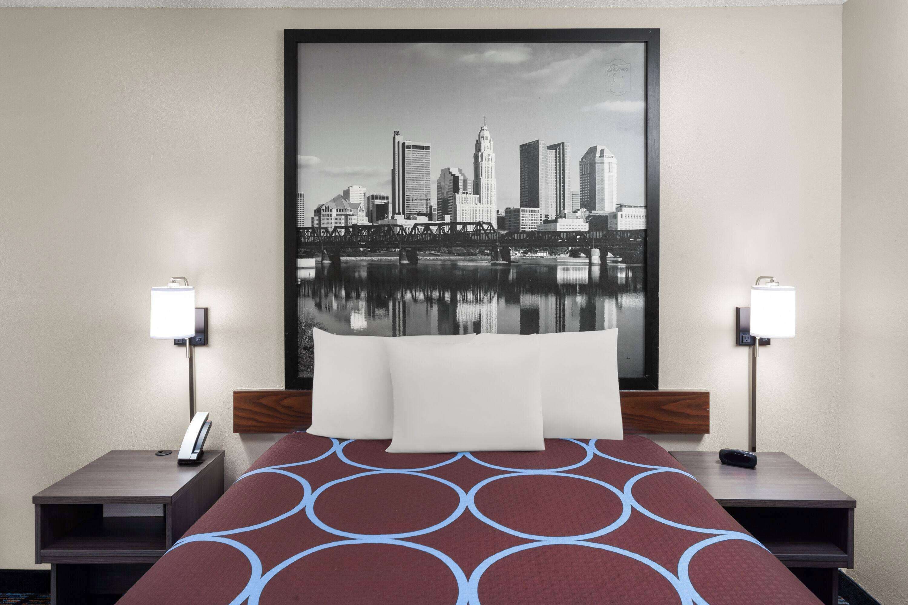 A hotel room with a large bed, two bedside tables, a cityscape artwork on the wall, and a nightstand with a lamp.