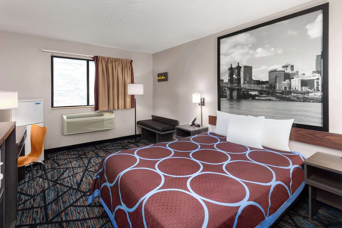 Een hotelkamer met een groot bed, een bureau, een stoel, een nachtkastje, een lamp, een raam met gordijnen, een airconditioning en uitzicht op een stadsbeeld.