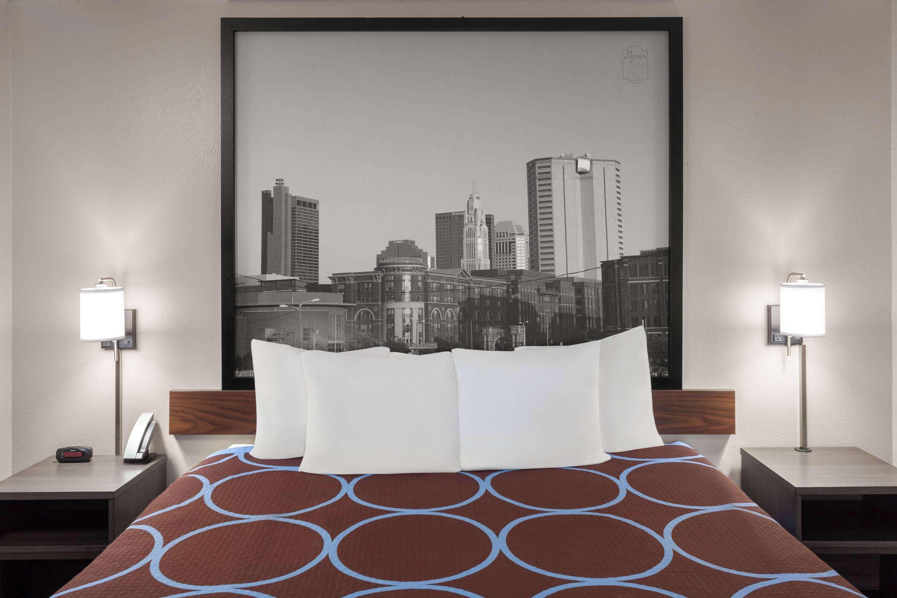 Une chambre d’hôtel avec un lit, deux tables de chevet et une œuvre d’art représentant un paysage urbain accrochée au mur.