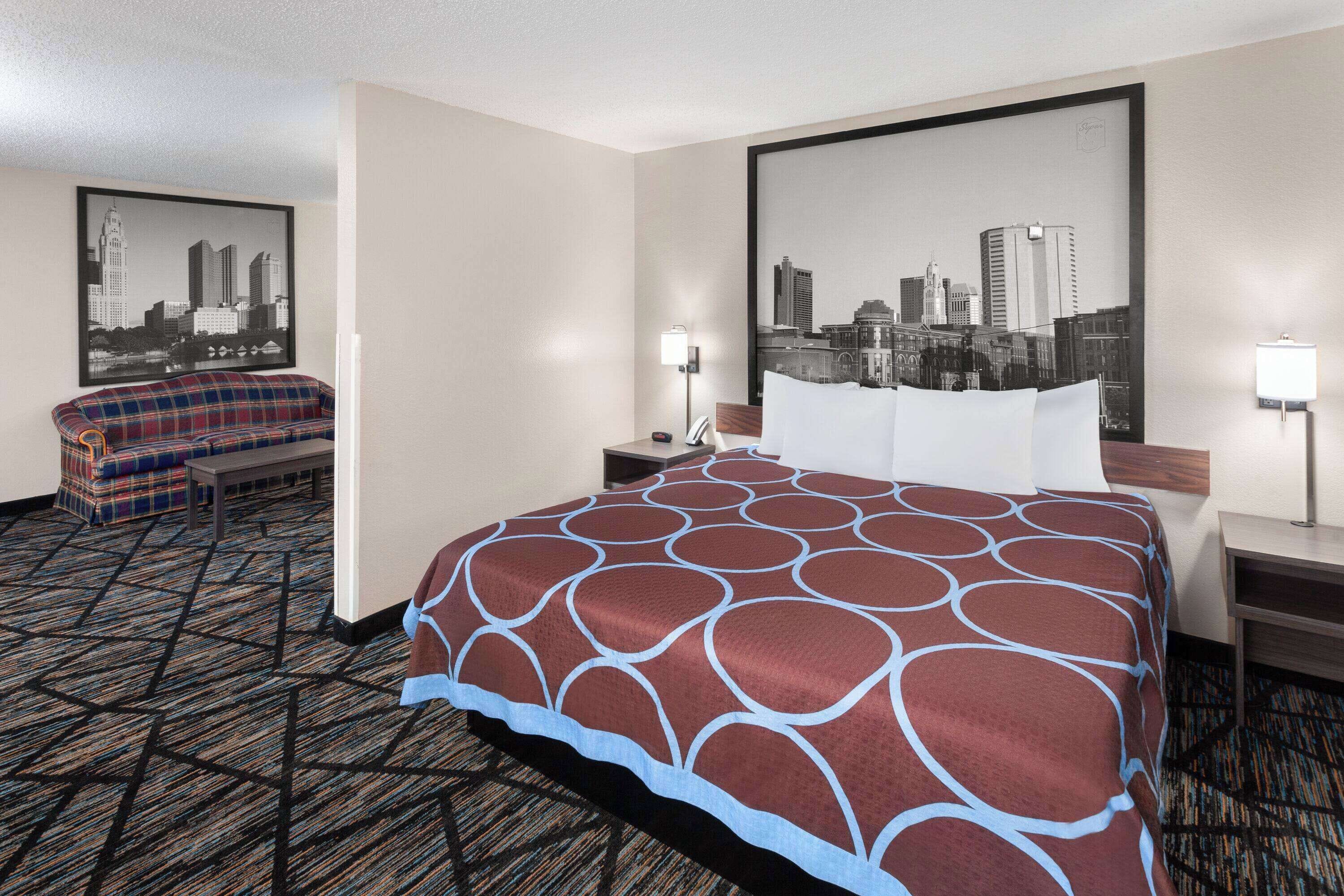 A hotel room with a large bed, a sofa, two nightstands, and a cityscape painting on the wall.