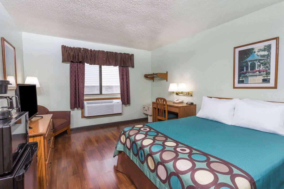 A hotel room with a bed, a desk with a chair, a lamp, a picture on the wall, and an air conditioning unit.