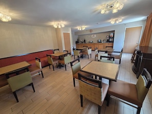Interior de un restaurante con varias mesas y sillas, una zona de cocina al fondo y un techo con luces colgantes.
