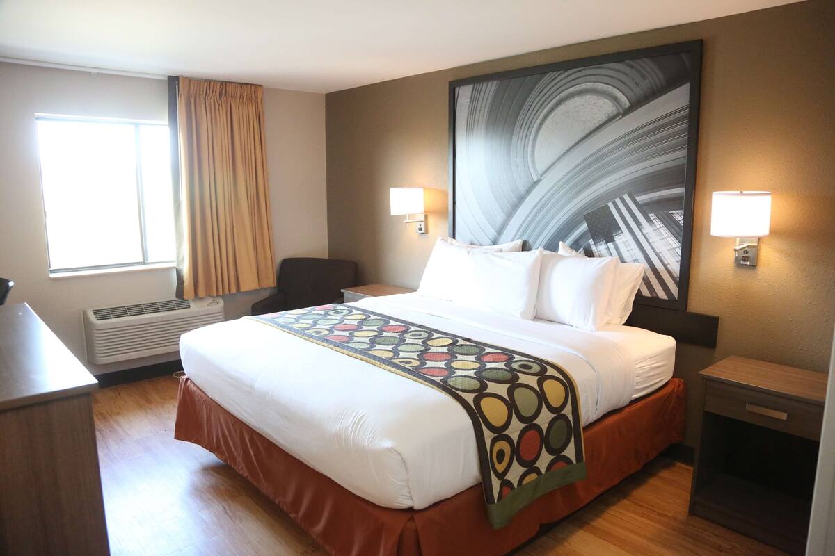 Una habitación de hotel con una cama grande, dos lámparas de noche, un cuadro abstracto grande en la pared, una ventana con cortinas y un escritorio.