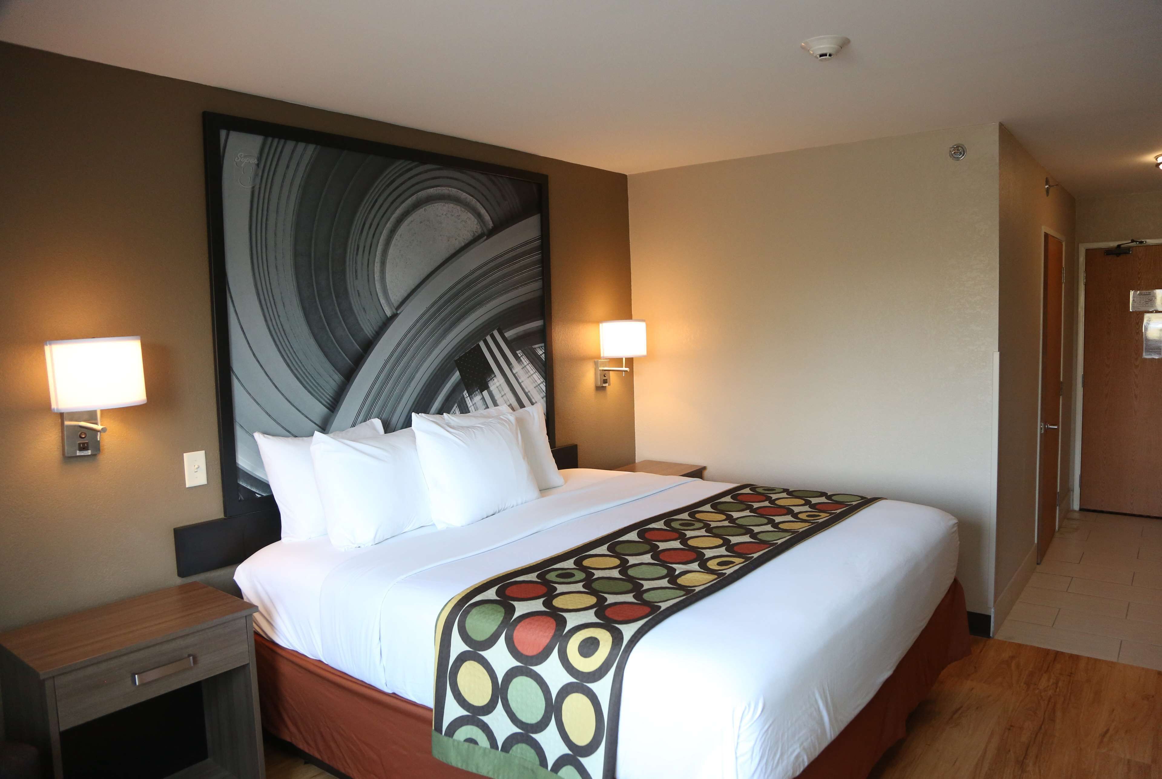 A hotel room with a large bed, two bedside lamps, a headboard with a large abstract artwork, and a wooden floor.