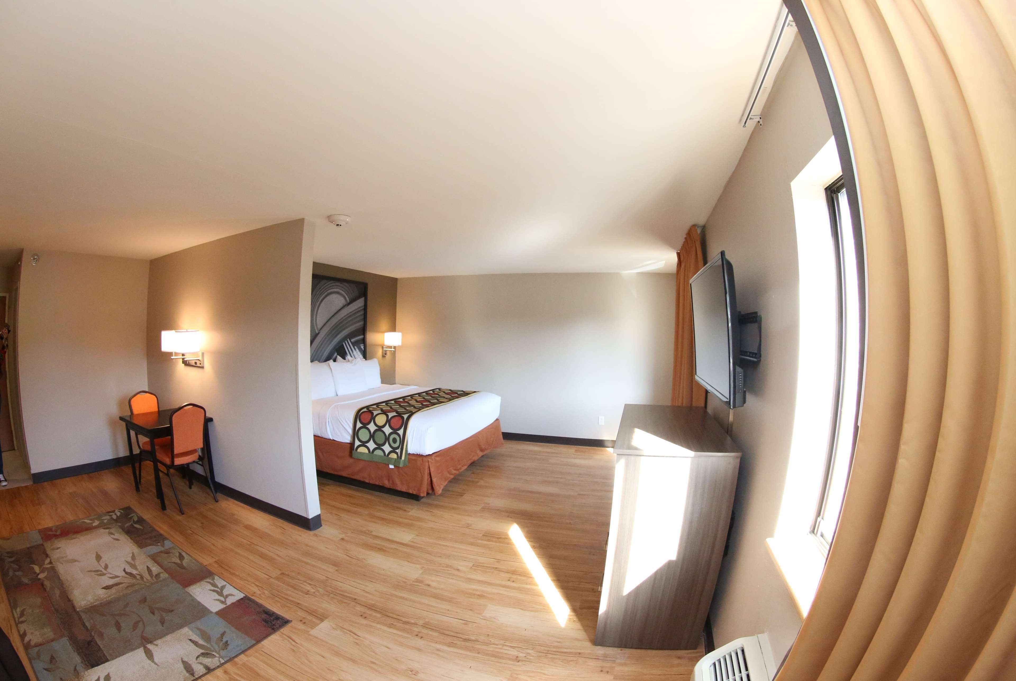A hotel room with a bed, a desk with a chair, a TV mounted on the wall, and a large window with curtains.