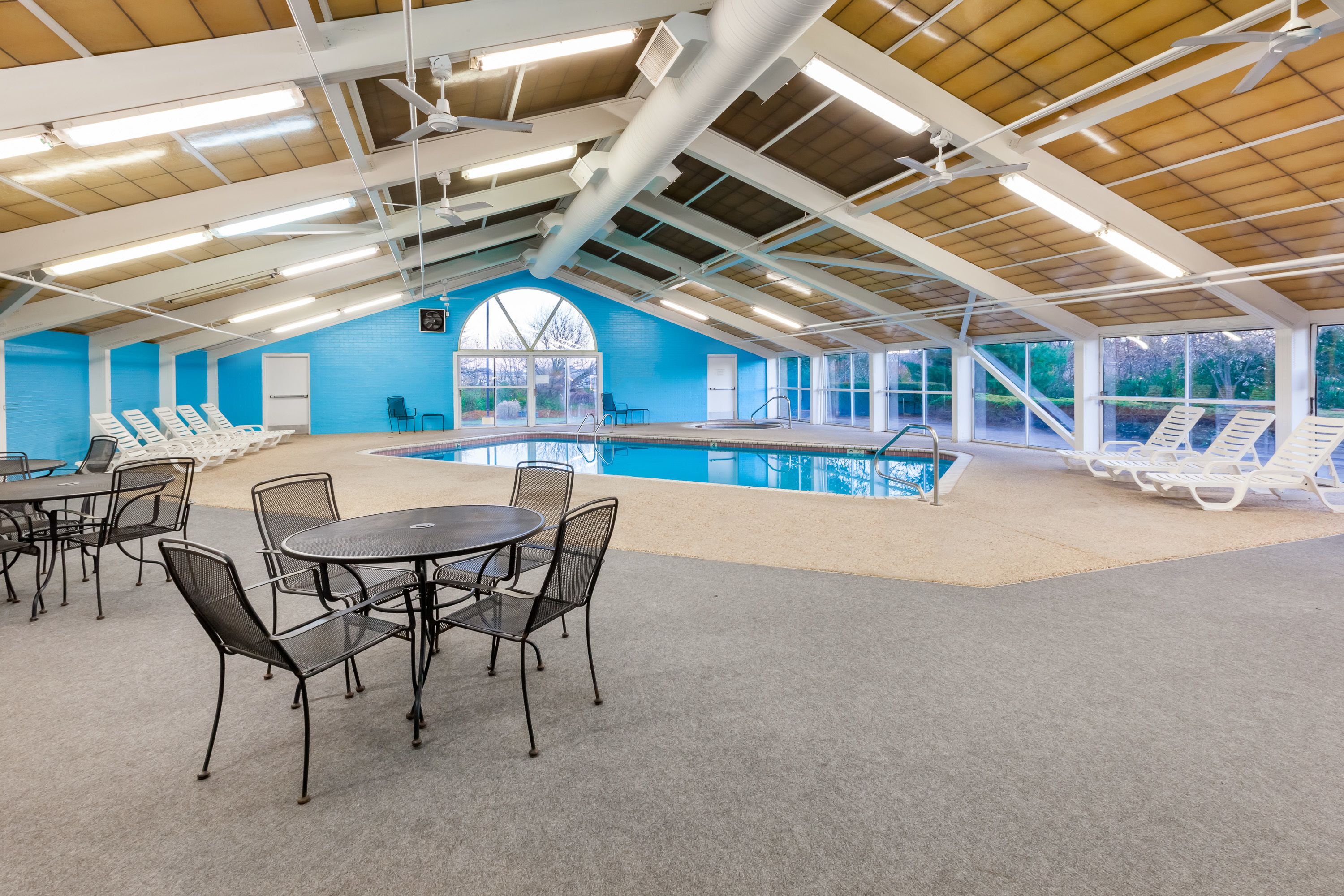 An indoor swimming pool with lounge chairs and a table, surrounded by large windows and a high ceiling with exposed beams.