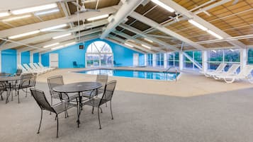 An indoor swimming pool with lounge chairs and a table, surrounded by large windows and a high ceiling with exposed beams.