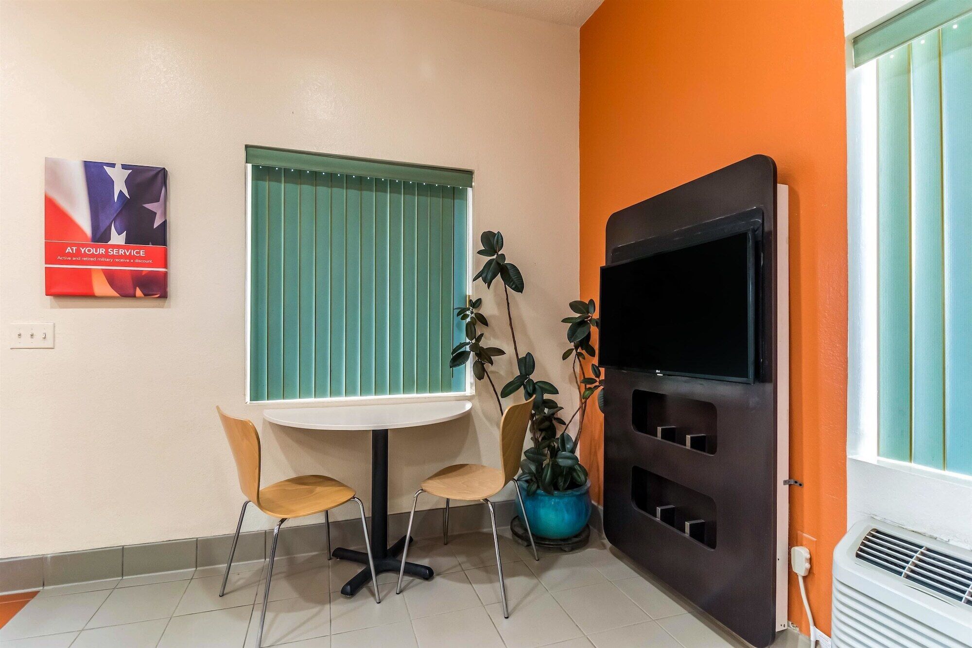 Une petite pièce avec une fenêtre de couleur turquoise, un téléviseur noir fixé au mur, une petite table avec deux chaises, une plante en pot et une affiche accrochée au mur.