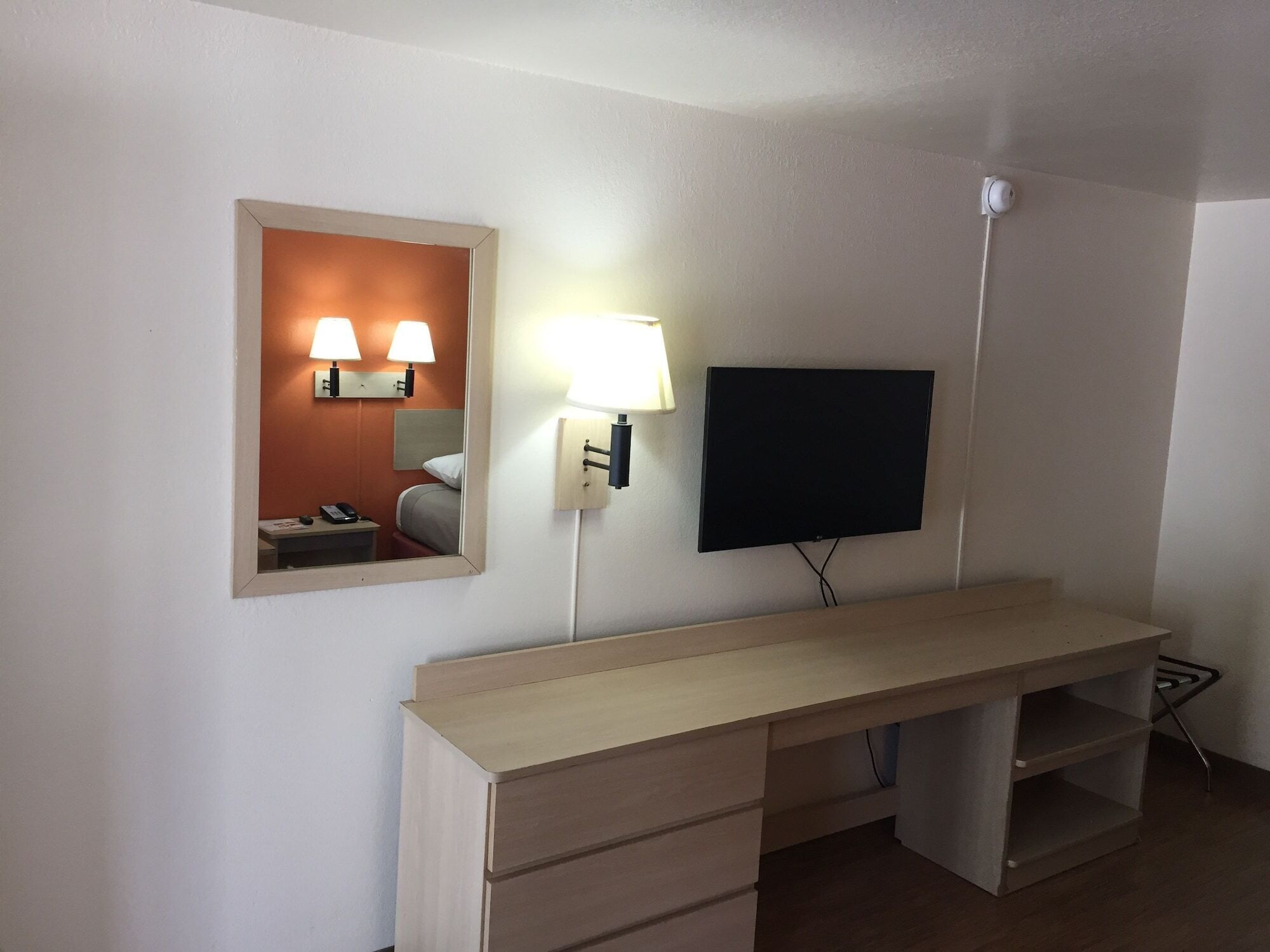 A hotel room with a wooden desk, a flat-screen TV mounted on the wall, a bedside lamp, and a mirror reflecting the room's interior.