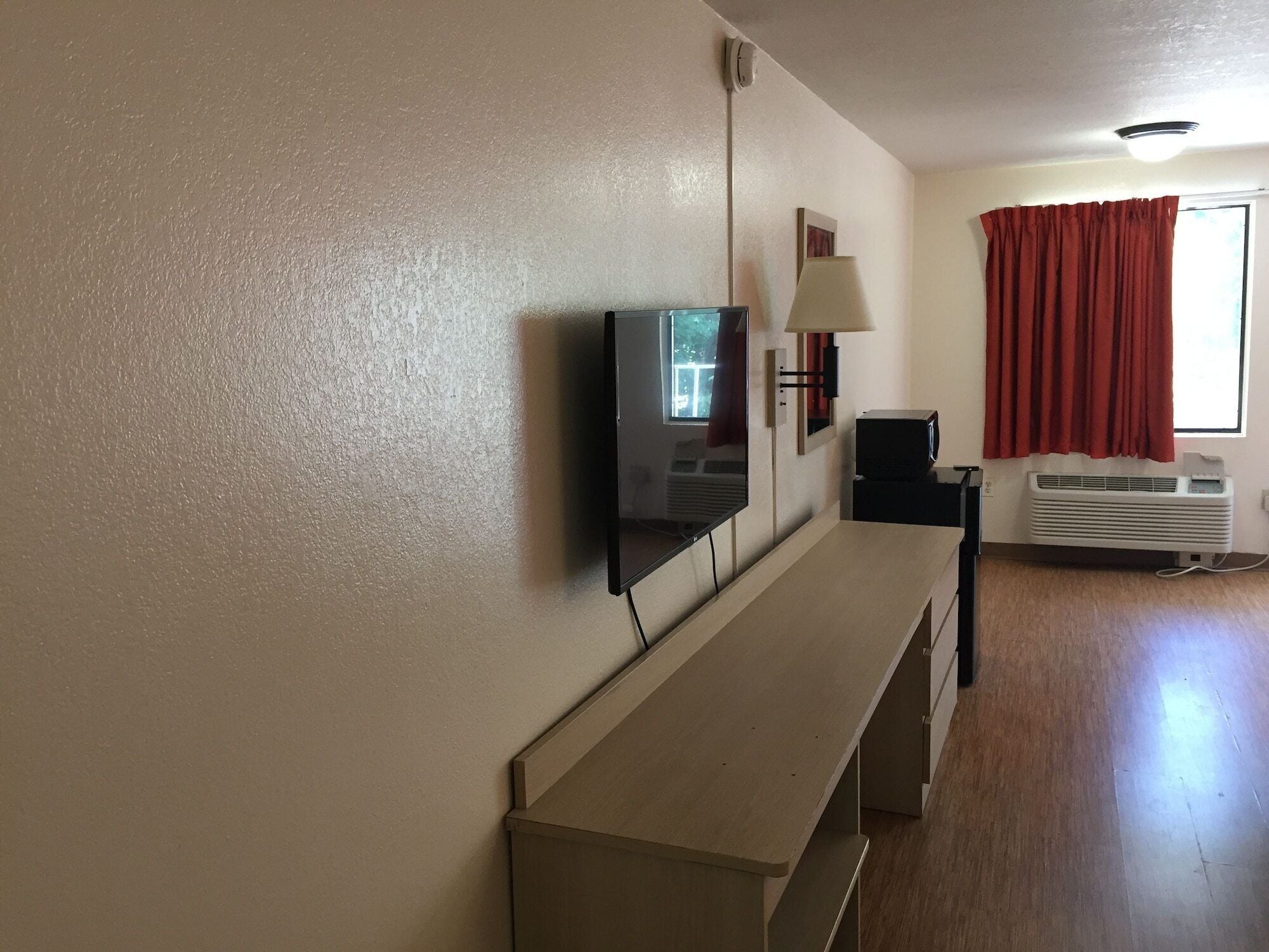 A room with a wooden bench, a television mounted on the wall, a lamp, and a window with red curtains.