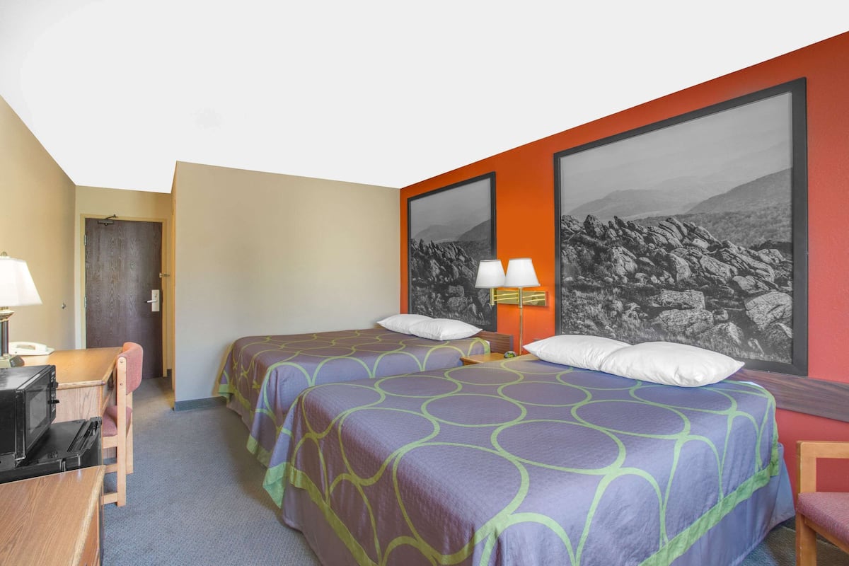 Ein Hotelzimmer mit zwei Betten, einem Schreibtisch, einem Stuhl, einer Lampe und einem großen, gerahmten Bild an der Wand.
