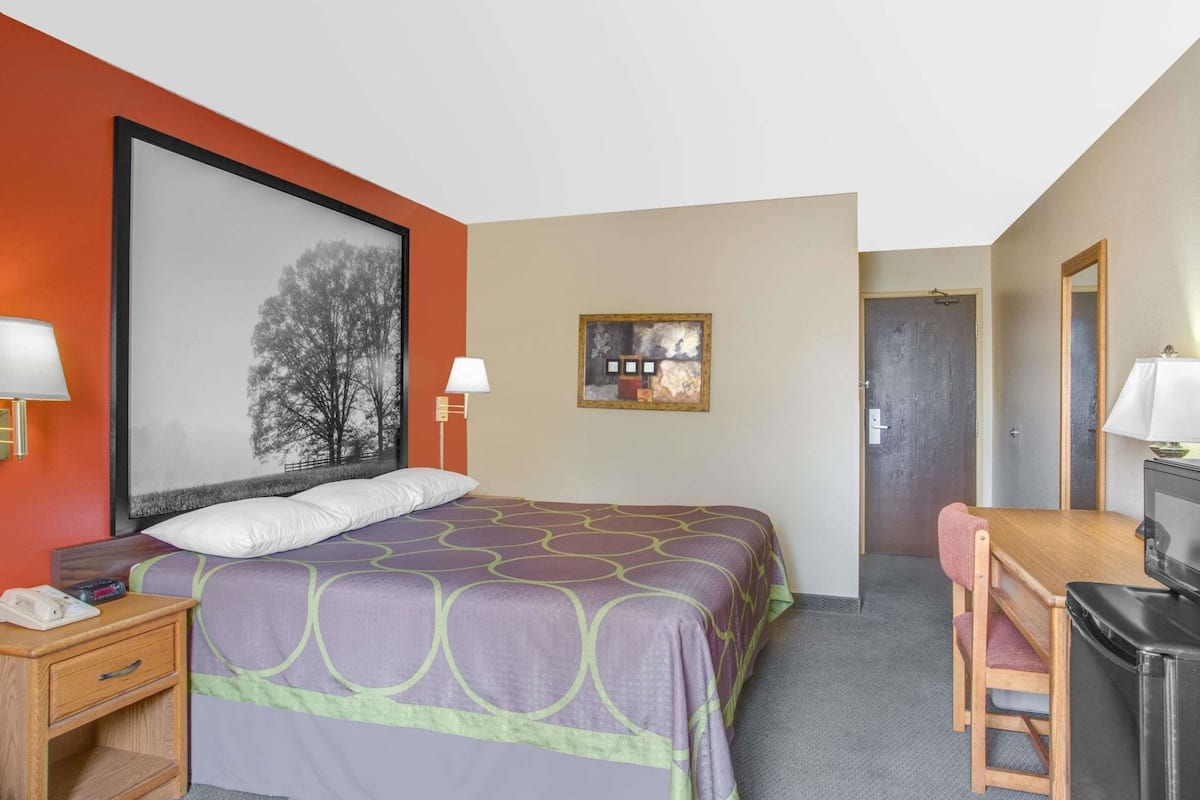 Ein Hotelzimmer mit Bett, Nachttisch, Schreibtisch, Stuhl, Fernseher und Kunstwerken an den Wänden.