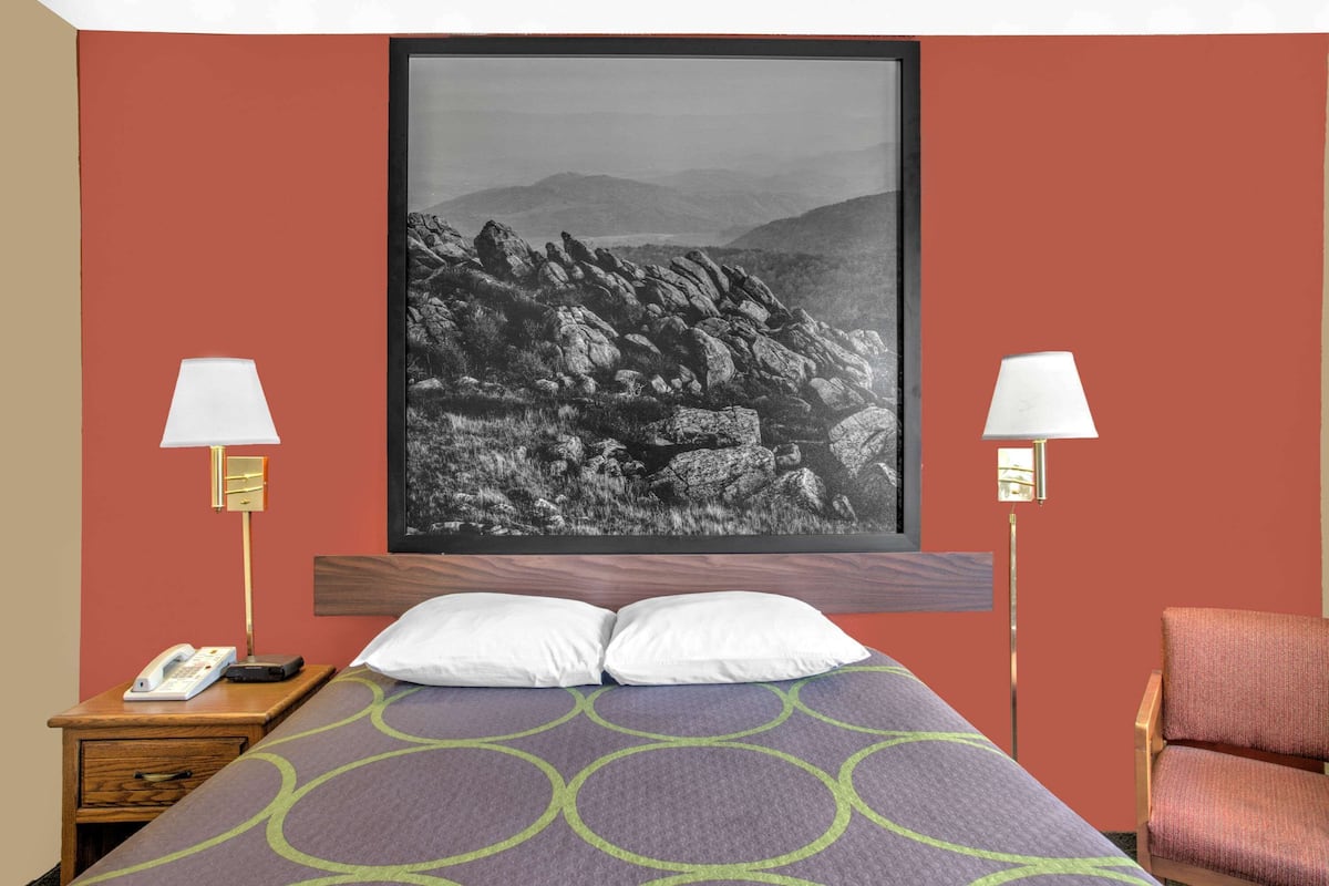 Ein Hotelzimmer mit einem Bett, zwei Nachttischlampen, einem Telefon, einem Sessel und einem großen gerahmten Bild an der Wand.