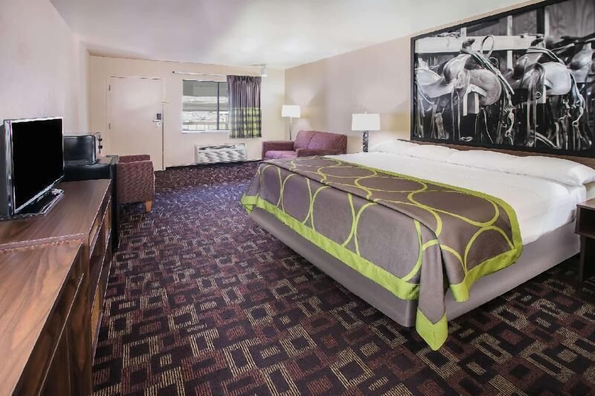 A hotel room with a bed, a desk with a TV, a chair, a window with curtains, and a painting on the wall.