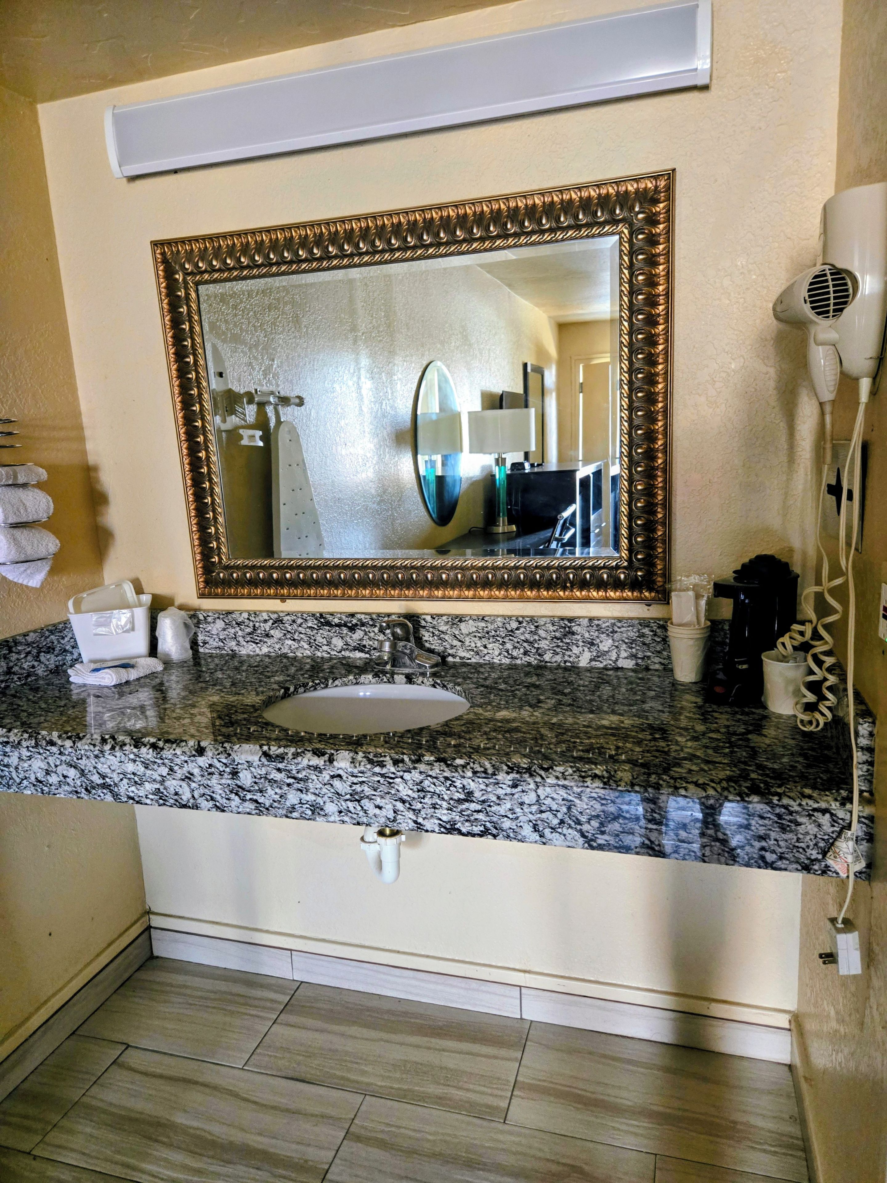 Un baño con encimera de granito, lavabo, espejo con marco decorativo, secador de pelo y toallero.