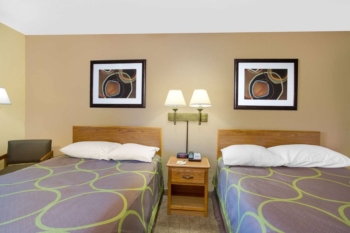 Habitación de hotel con dos camas, cada una con cabecero de madera, mesita de noche con lámpara y cuadros en la pared.