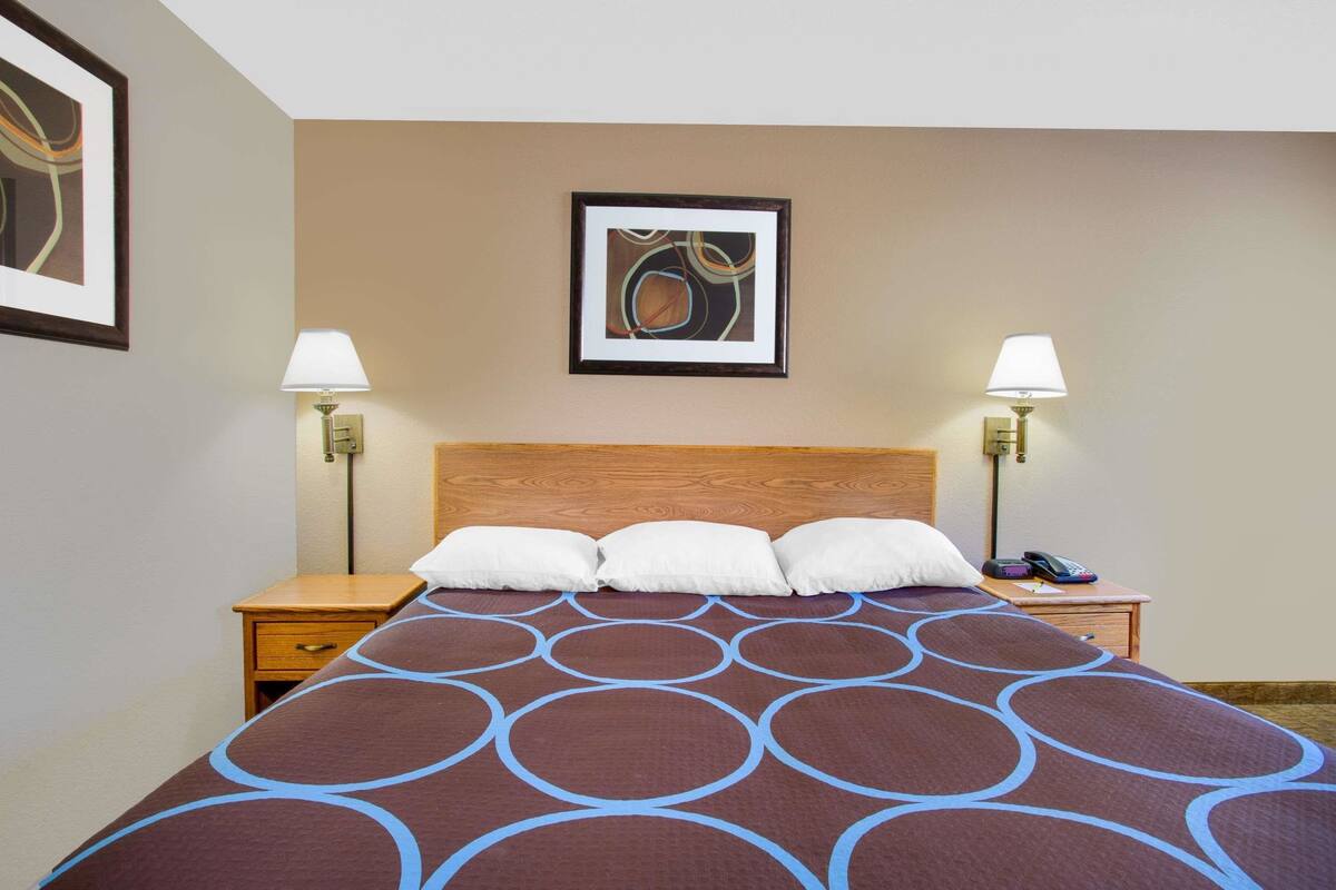 Habitación de hotel con una cama grande, dos mesitas de noche, una obra de arte enmarcada sobre la cama y dos lámparas fijadas a la pared.