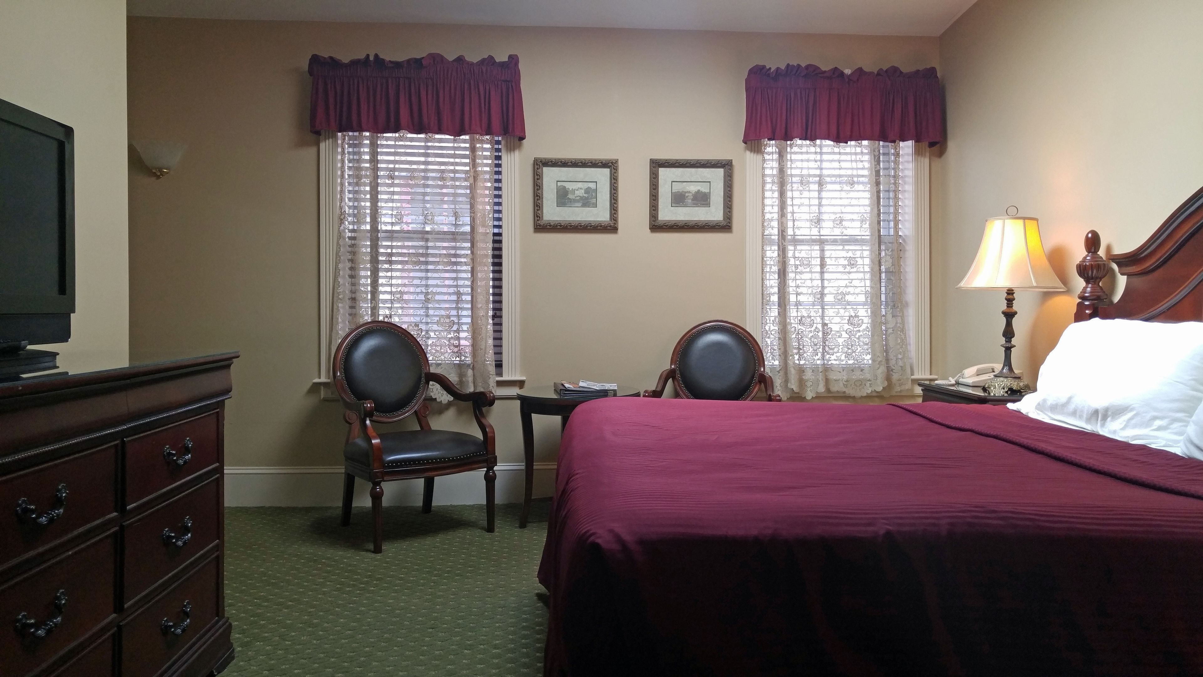 Habitación de hotel con cama, dos sillones, cómoda, mesita de noche con lámpara y dos ventanas con cortinas.