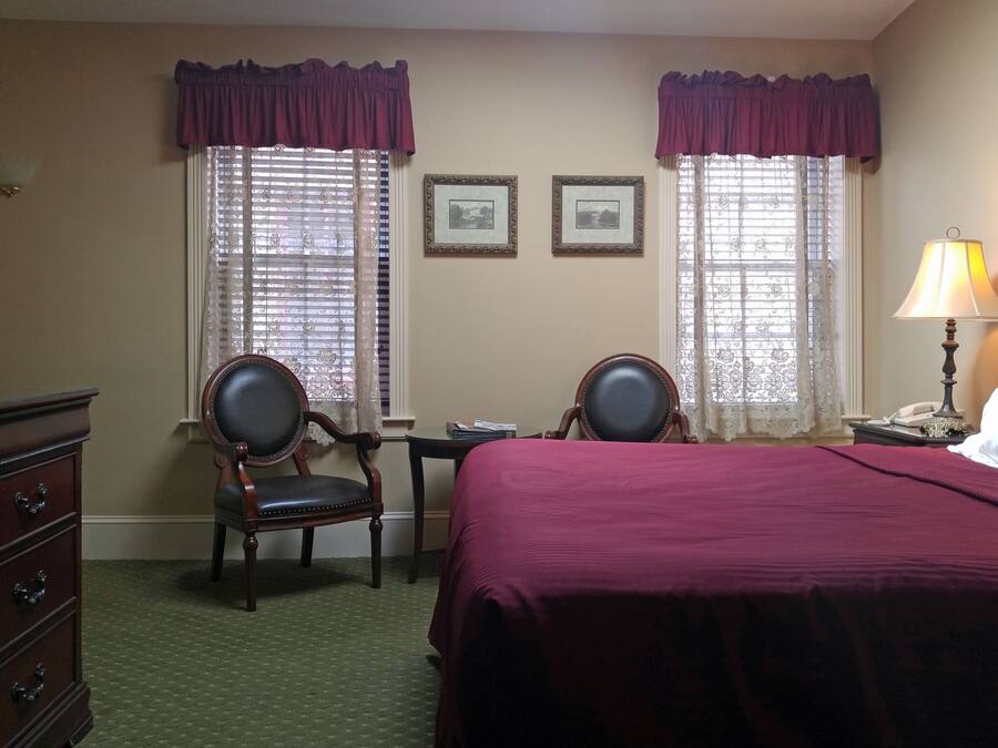 A hotel room with a bed, two chairs, a dresser, a nightstand with a lamp, and two windows with curtains.