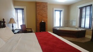 A hotel room with a large bed, a red throw, a wooden headboard, a desk with a lamp, a chair, a brick wall, and a bathroom with a bathtub and window blinds. - Inn at Jim Thorpe (Jim Thorpe)