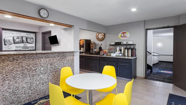 Un hall d’hôtel avec un comptoir d’accueil, une horloge, un téléviseur fixé au mur, un buffet pour le petit-déjeuner et des chaises jaunes disposées autour d’une table.
