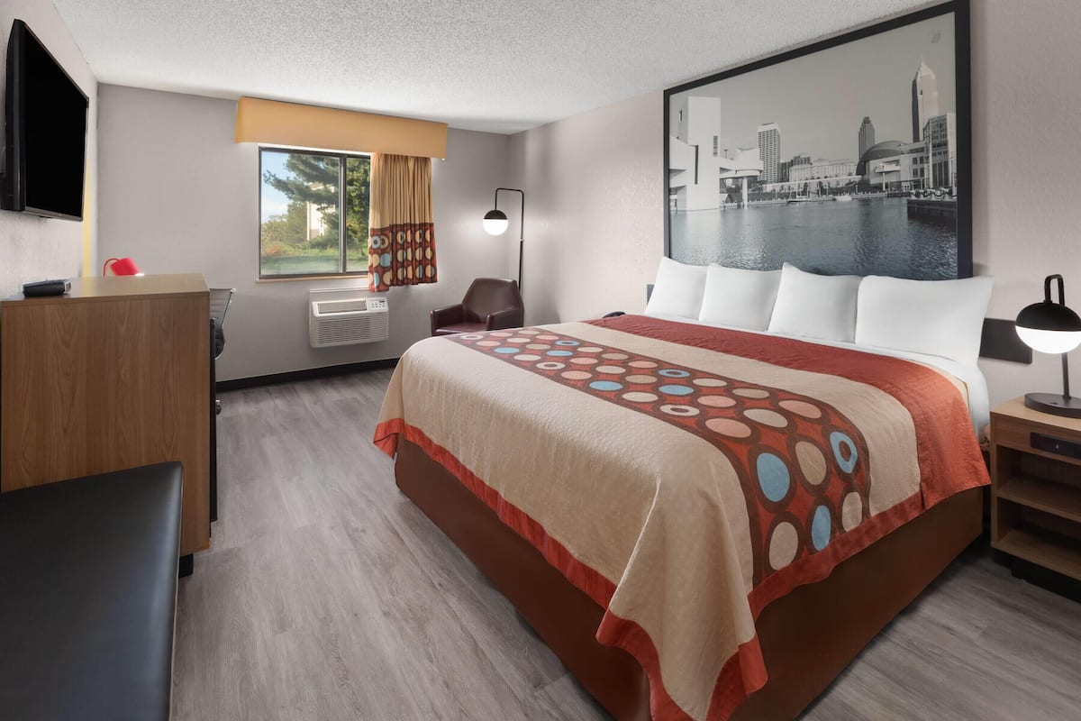 A hotel room with a bed, a desk, a chair, a TV, a window with curtains, an air conditioning unit, a lamp, and a cityscape painting on the wall.