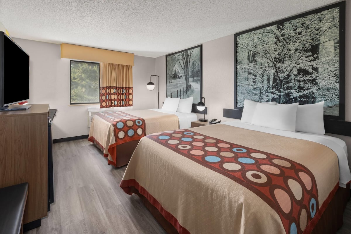 A hotel room with two beds, a desk, a chair, a TV, and large windows with curtains.