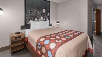Quarto de hotel com uma cama grande, uma mesa de cabeceira com luminária, uma janela com vista para árvores e uma porta.