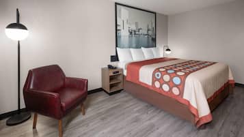 Habitació d'hotel amb un llit, un butacó vermell, una tauleta de nit, una làmpada i un quadre de paisatge urbà a la paret.