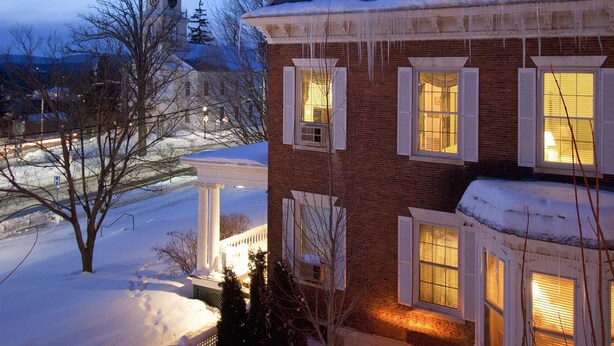 A snow-covered house with a brick exterior and white shutters, a snow-covered yard, and a church steeple in the background.