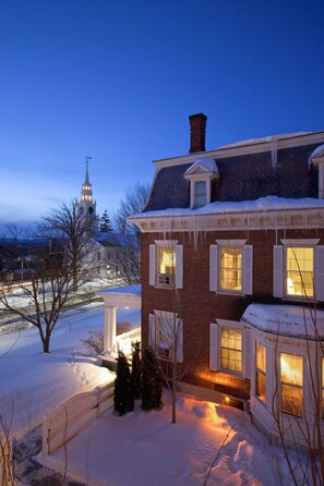 A snow-covered house with a brick exterior and white shutters, a snow-covered yard, and a church steeple in the background.