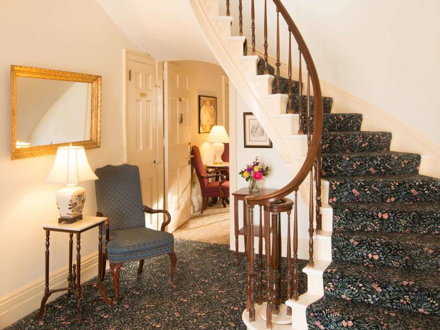 A grand staircase with a wooden handrail and white steps, leading to a room with a floral carpet, a chair, and a lamp.