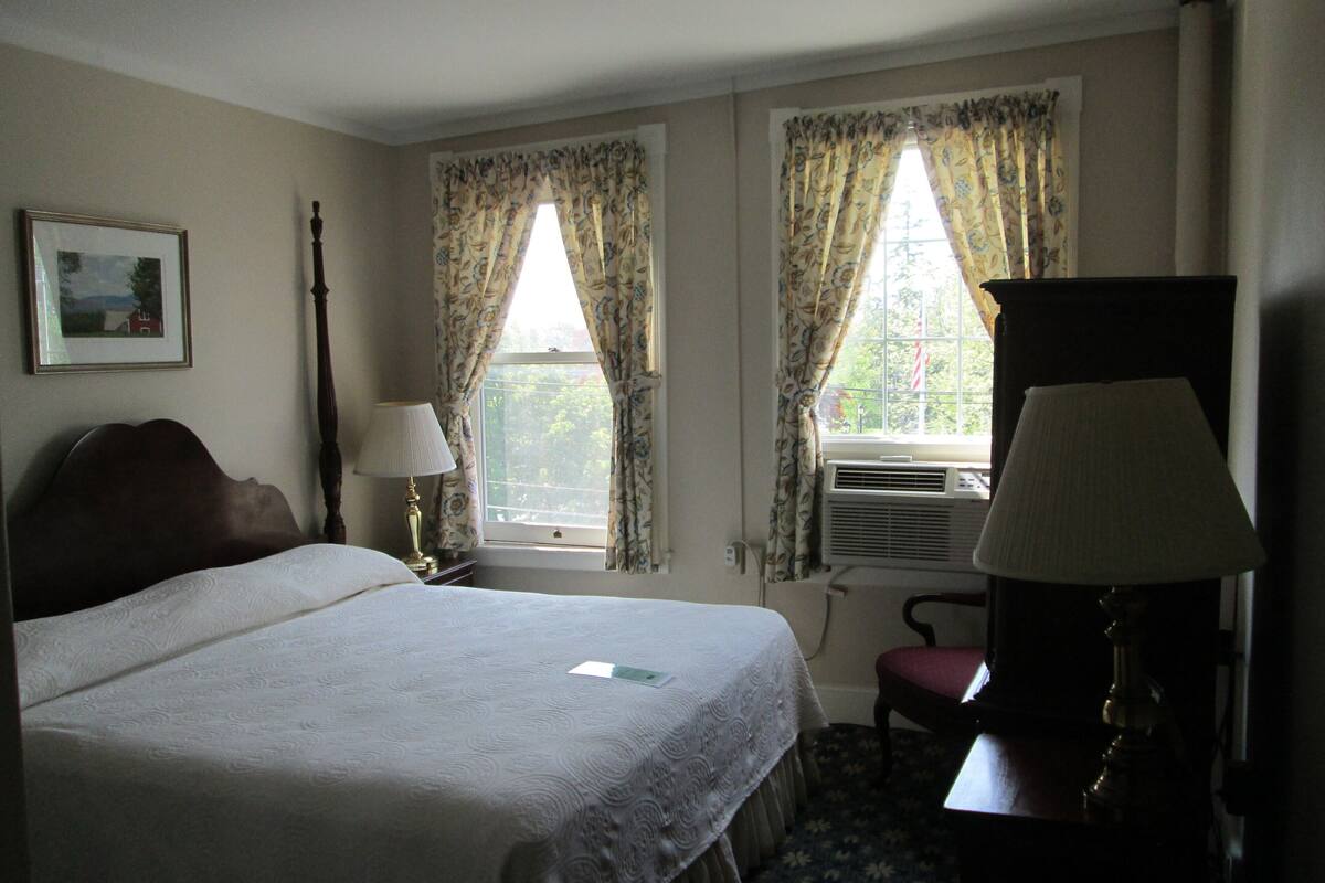 A hotel room with a bed, two windows with curtains, a desk with a lamp, and a chair.