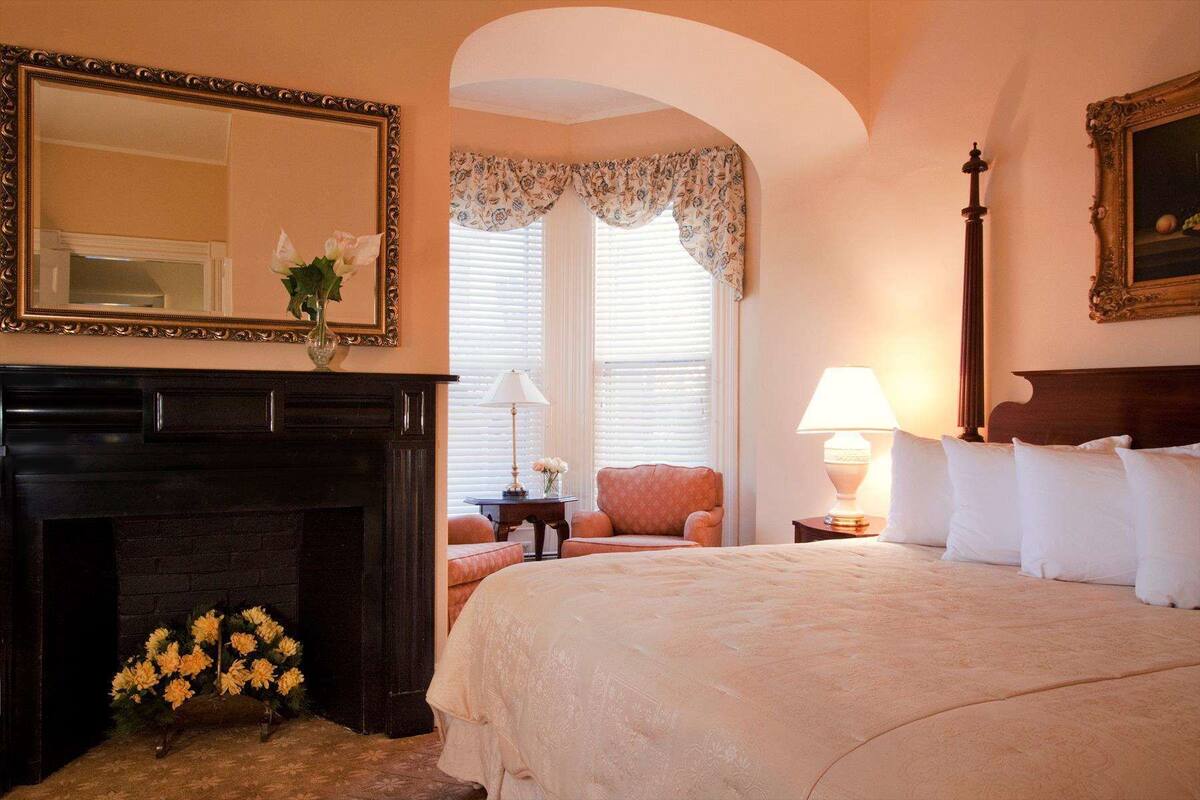A hotel room with a large bed, a fireplace, a mirror, and a window with floral curtains.