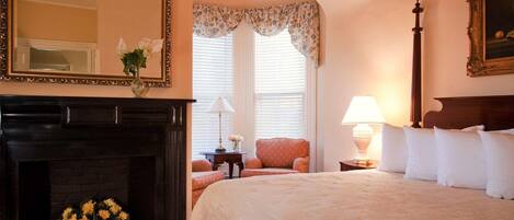 A hotel room with a large bed, a fireplace, a mirror, and a window with floral curtains.