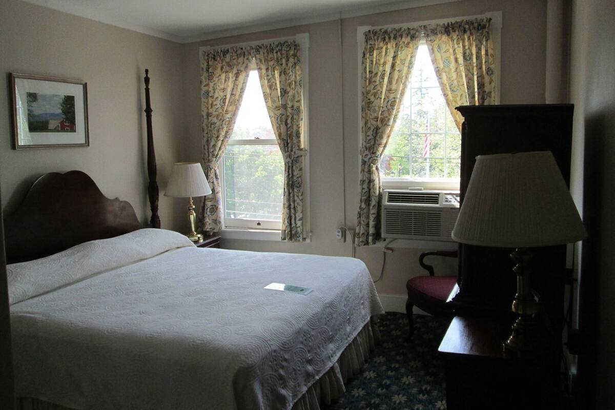 A hotel room with a bed, two windows with curtains, a desk with a lamp, and a chair.
