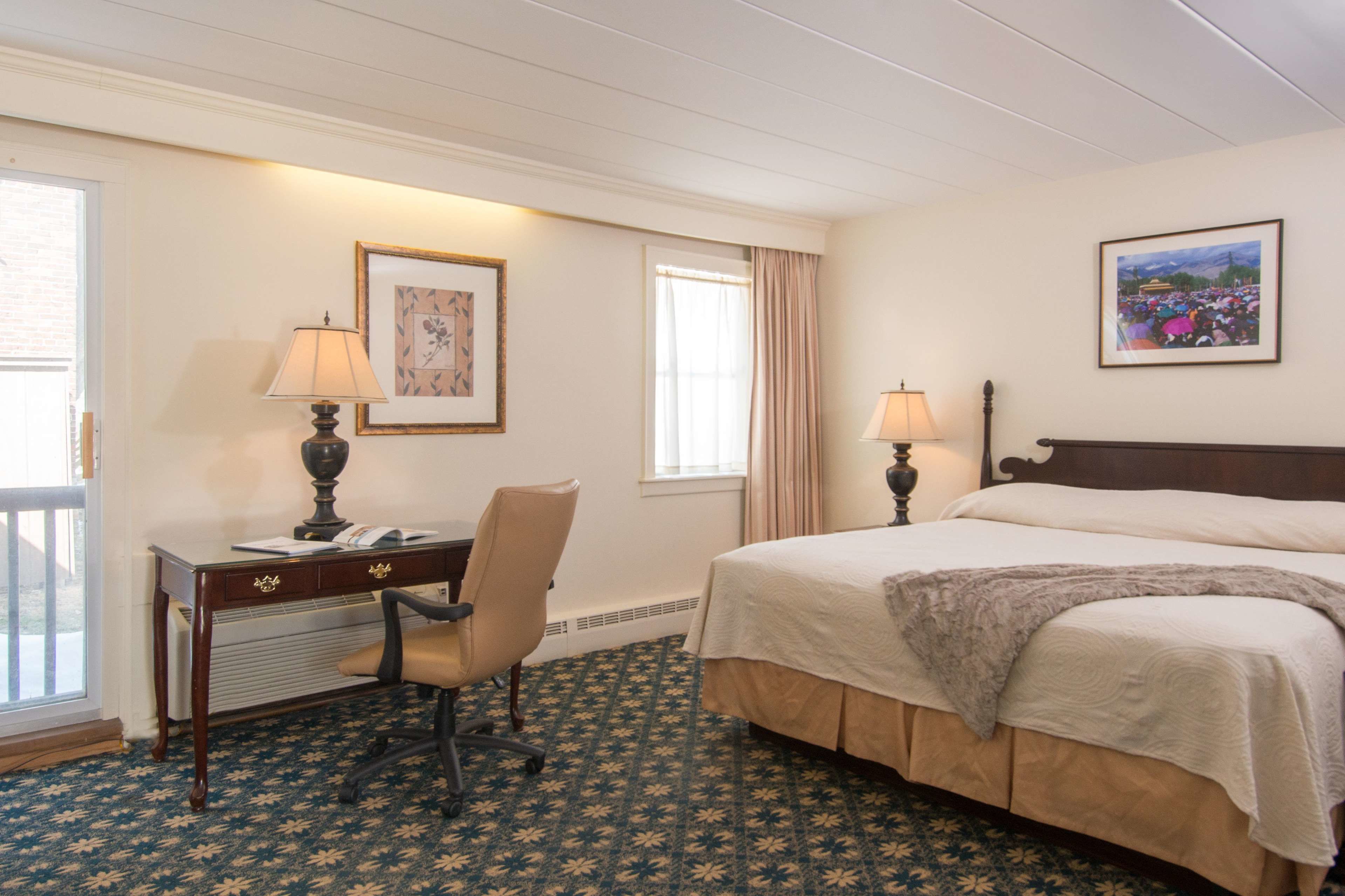 Habitación de hotel con cama, escritorio, silla, dos lámparas, un cuadro enmarcado y una ventana con cortinas.