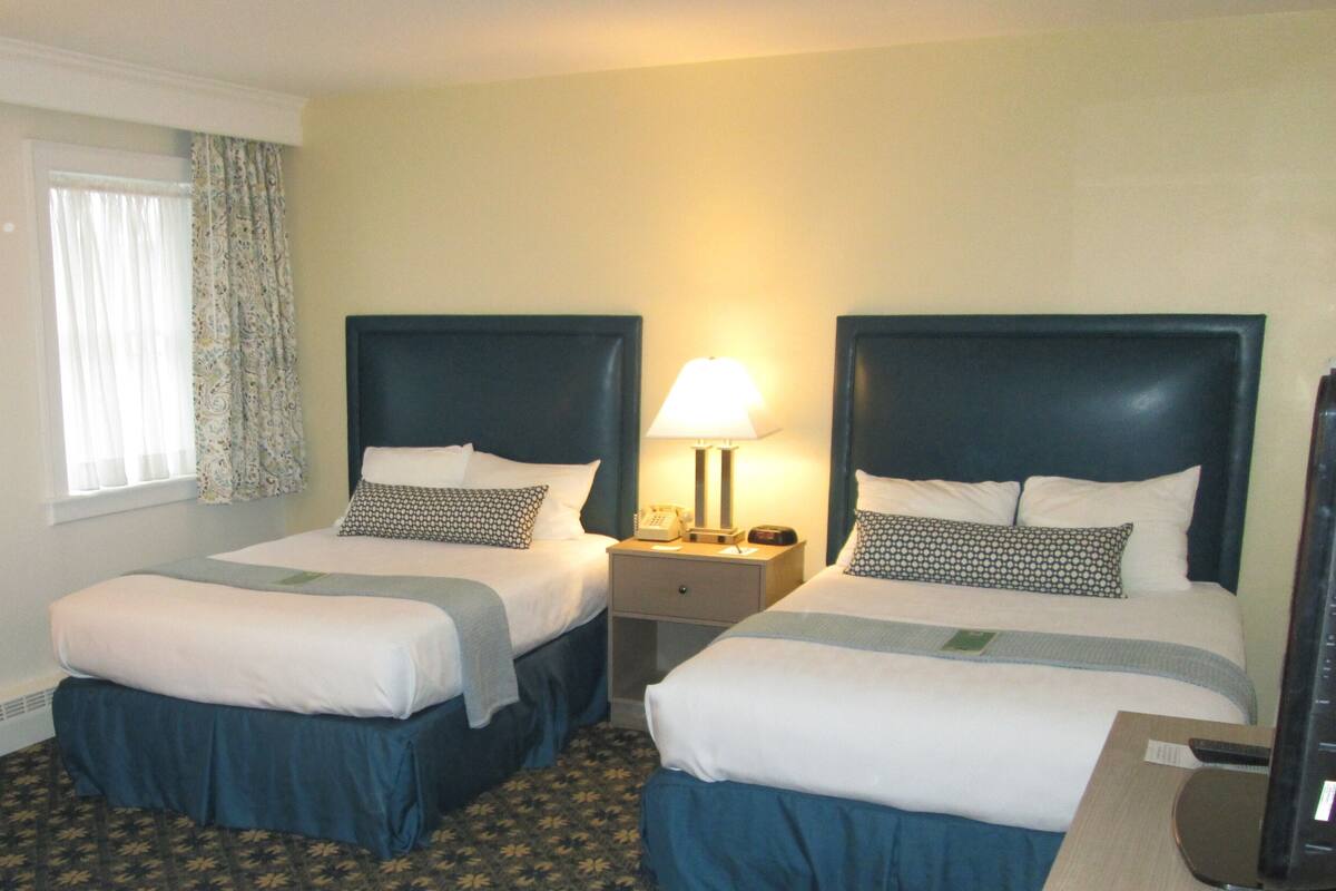 A hotel room with two beds, a nightstand with a lamp, and a window with curtains.