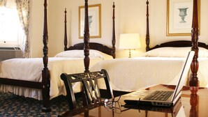 A four-poster bed with a laptop on a wooden desk, a chair, and a bedside table with a lamp.