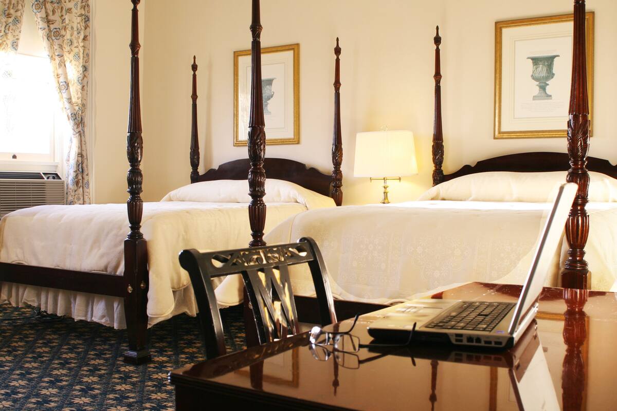 A four-poster bed with a laptop on a wooden desk, a chair, and a bedside table with a lamp.