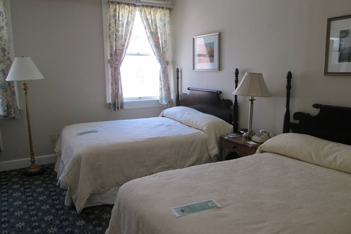 A hotel room with two single beds, a bedside table with a lamp, a framed picture on the wall, and a window with floral curtains.