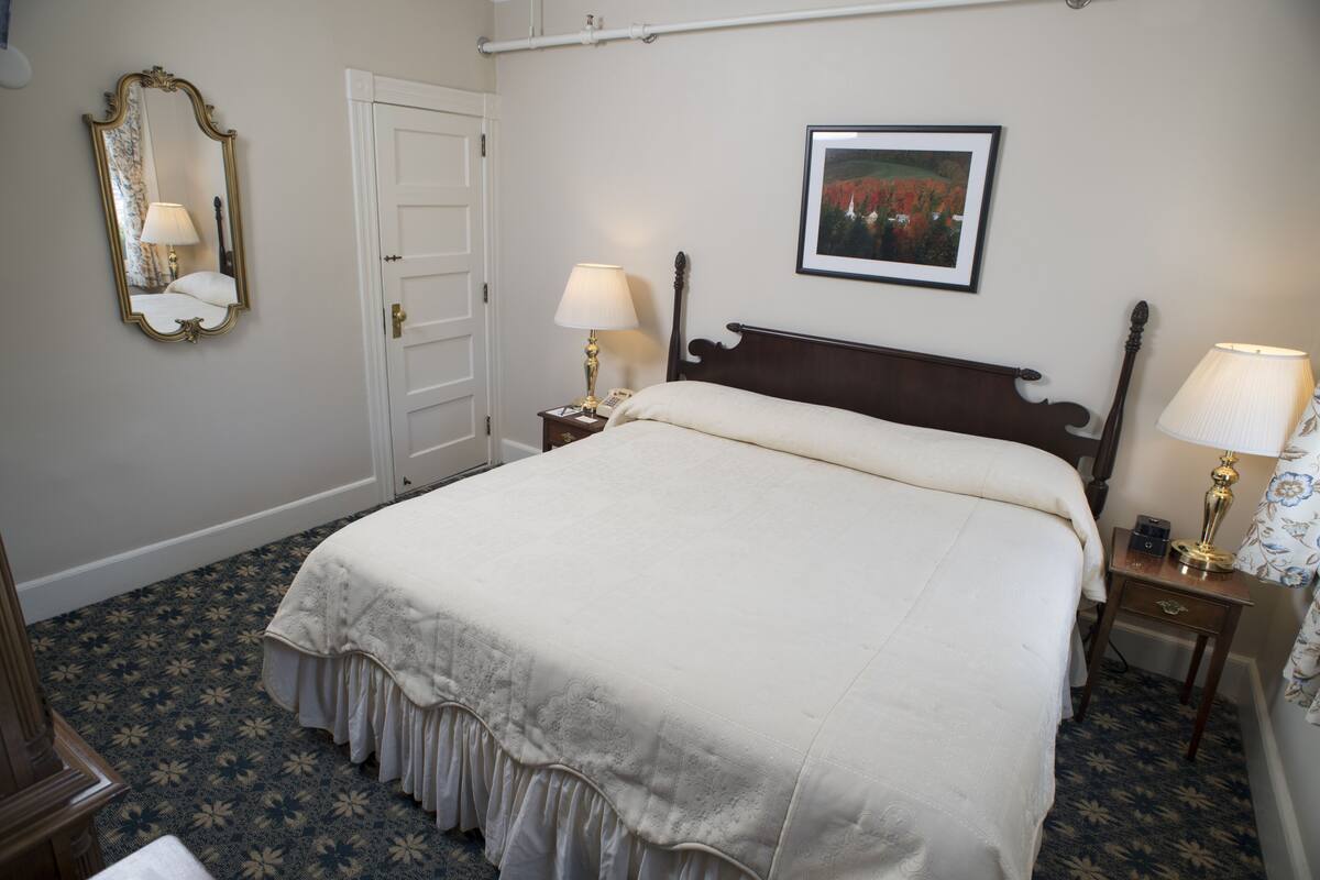 A neatly made bed with a white bedspread, two bedside tables with lamps, a mirror, a door, and a framed picture on the wall.